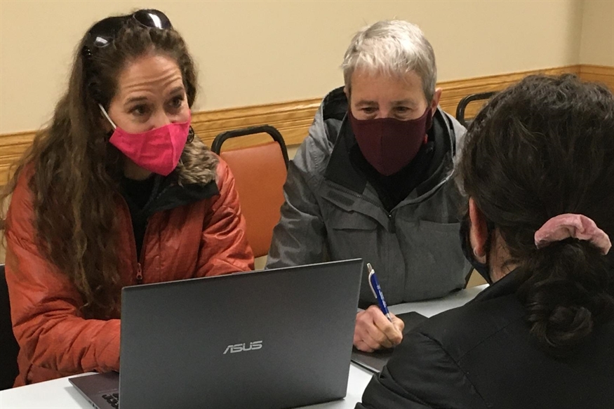 [ai] Two women wearing masks engage in a discussion at a table, looking at a laptop. One woman has curly brown hair and wears a pink mask, while the other has short gray hair and wears a dark mask. Another woman sits across from them, partially visible.